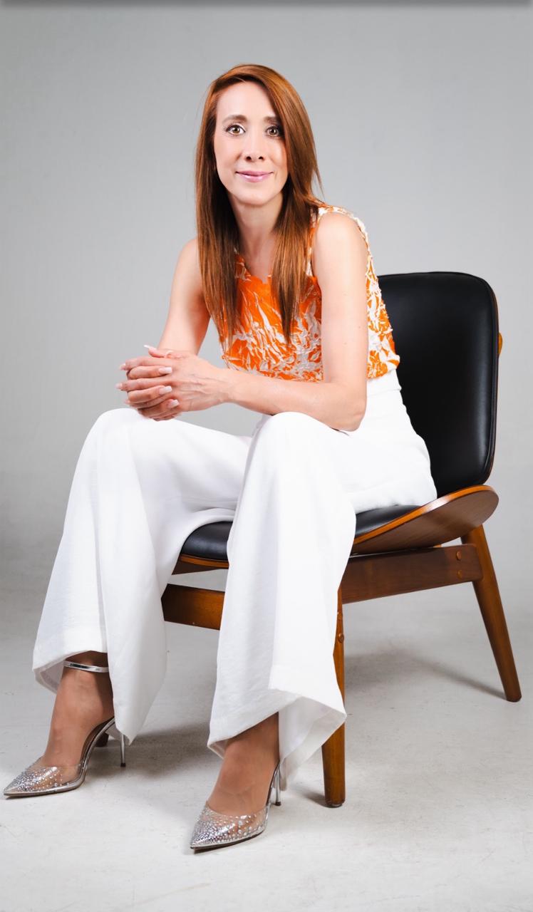 Lily Castellanos sentada, con blusa naranja y pantalón blanco